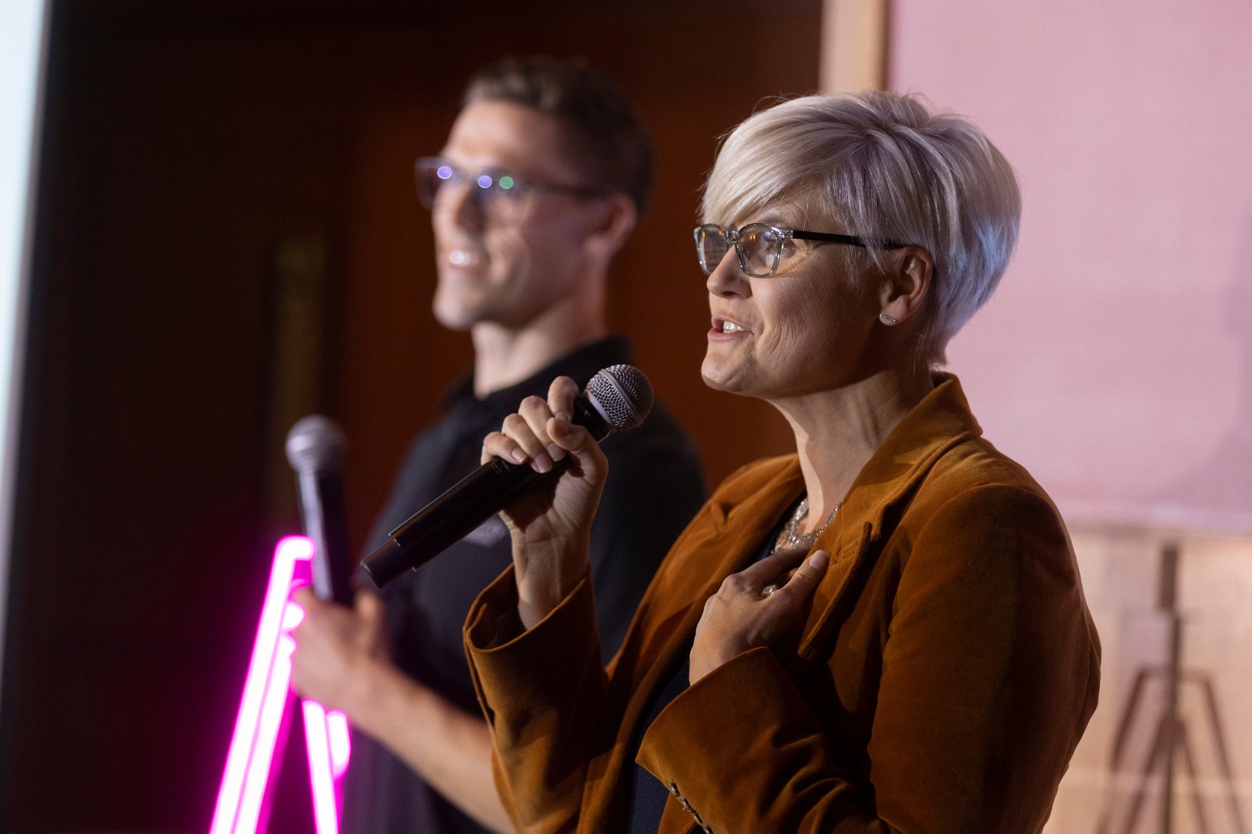 A woman with short gray hair and glasses holding a microphone, standing next to a man with glasses, also holding a microphone, in an indoor setting with warm lighting.