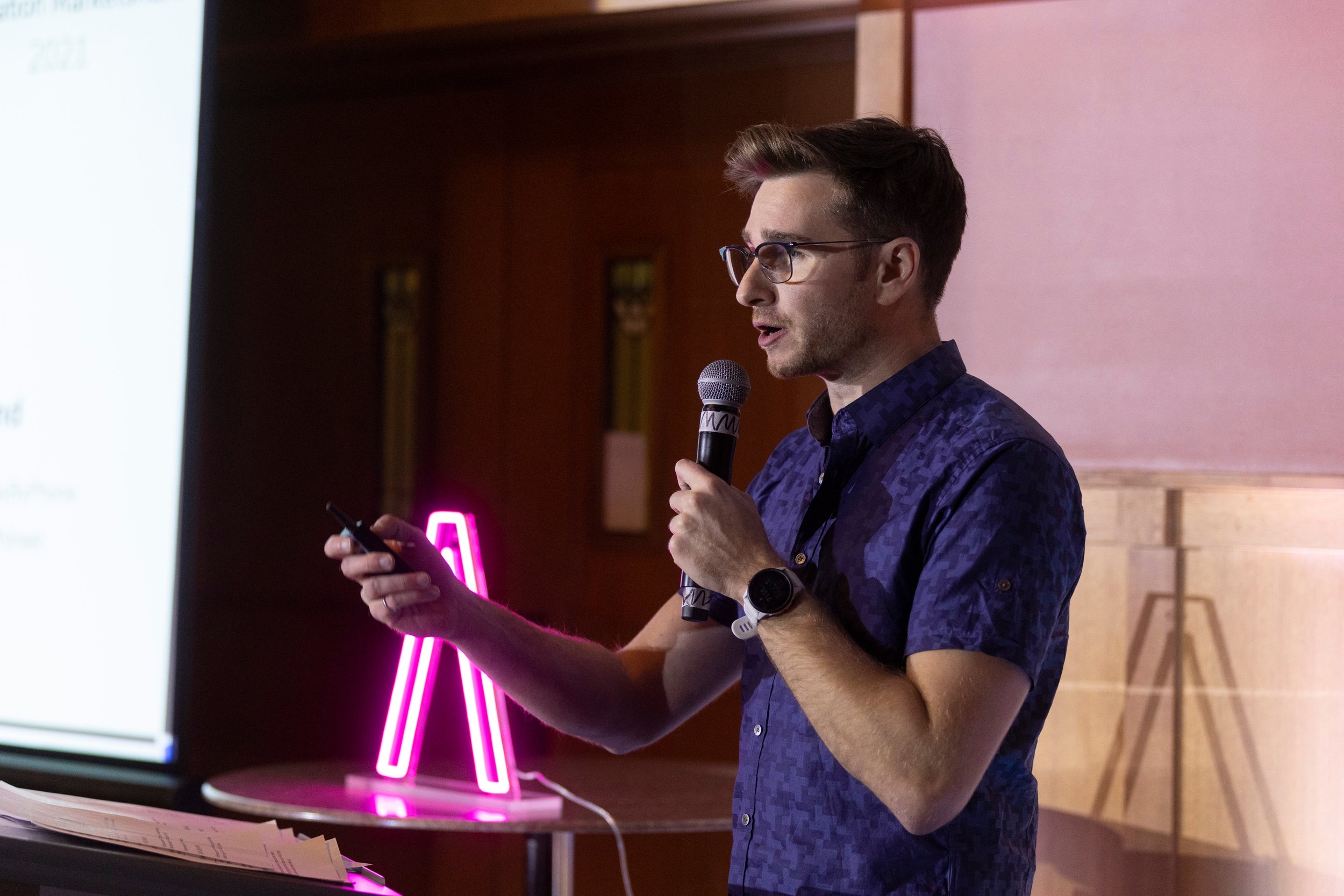 A man with glasses and a dark blue patterned shirt giving a presentation, holding a microphone in one hand and a remote in the other, with a pink neon letter 'A' in the background.