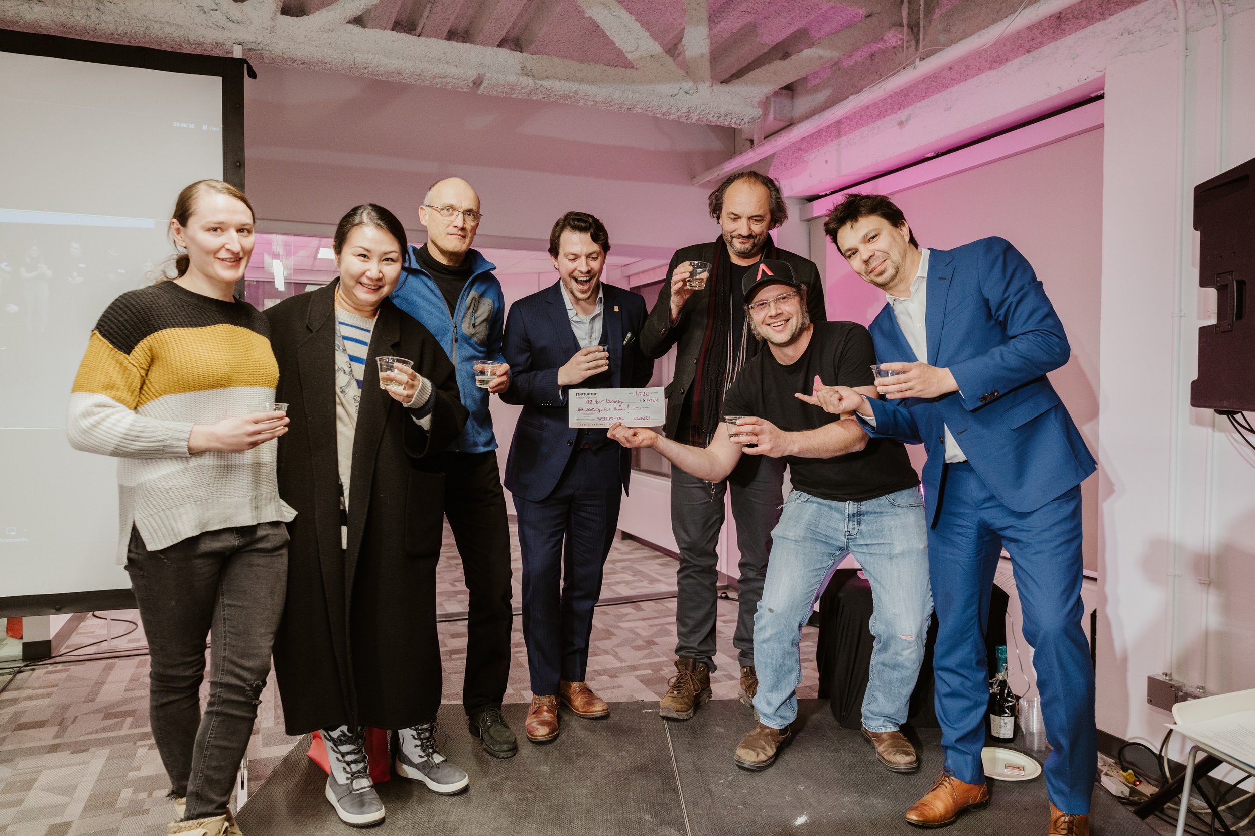 Group of people celebrating and holding a check in an indoor setting with pink lighting.