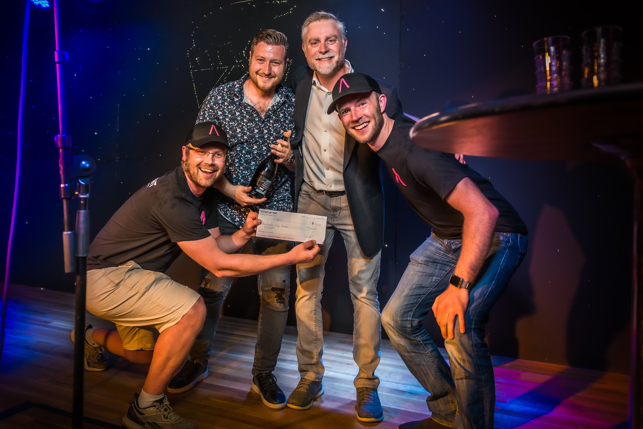 Group of five people celebrating, one holding a large check, one holding a bottle, in a dark room with colourful lights.