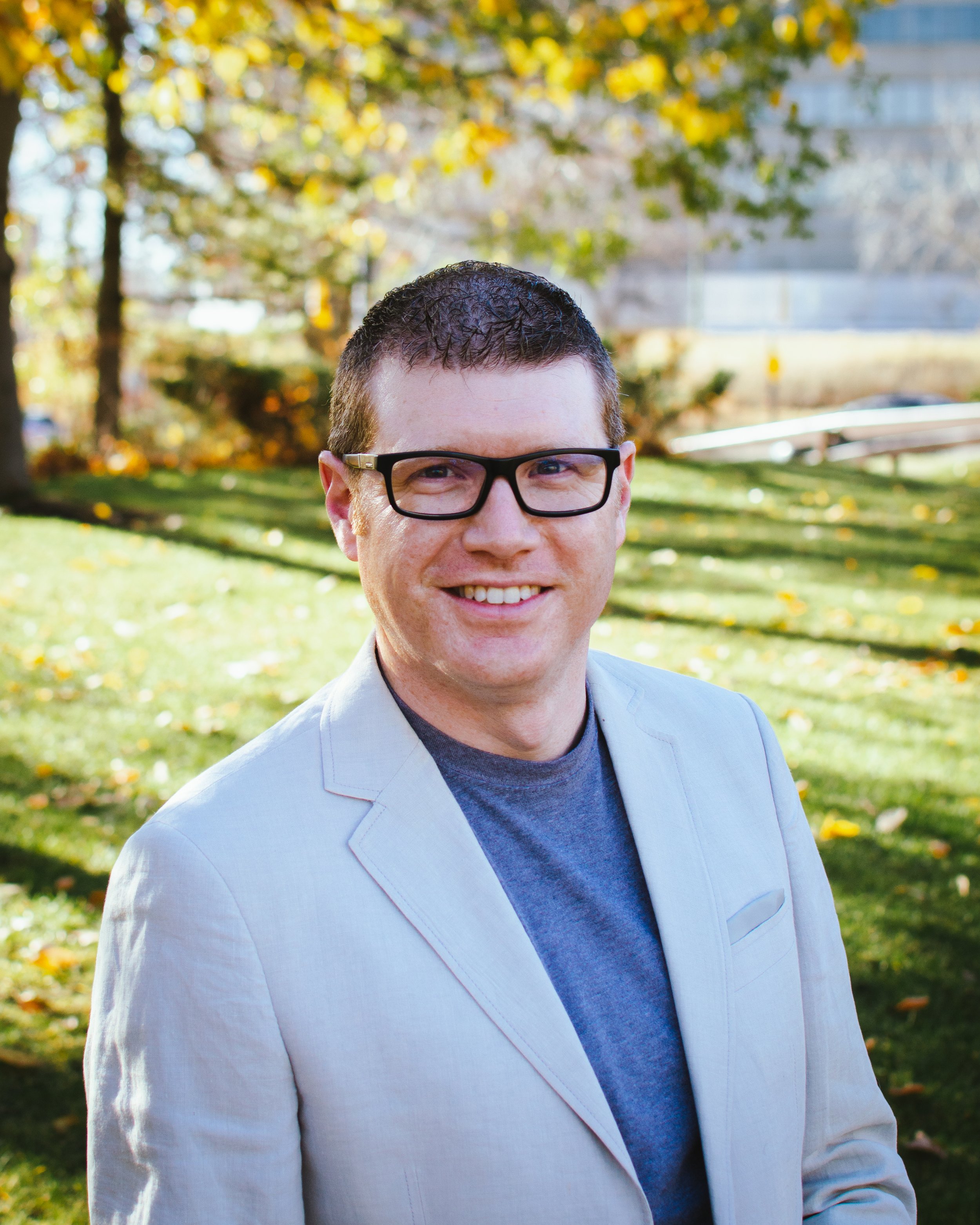A smiling man wearing glasses and a light-colored blazer outside in a park with trees and green grass in autumn.