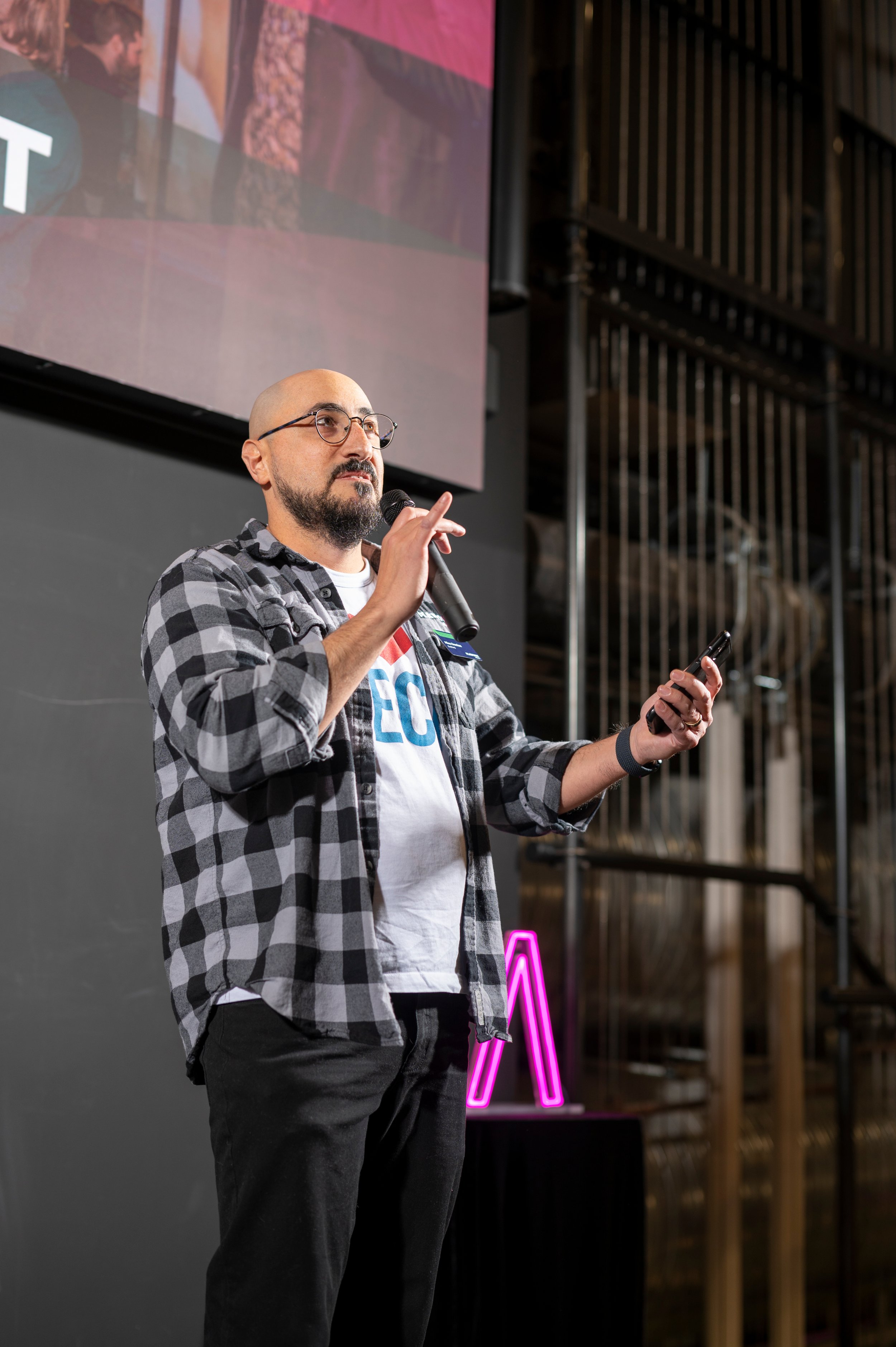 A man with glasses and a beard, wearing a plaid shirt over a T-shirt, holding a microphone in one hand and a remote in the other, standing in front of a large screen at pitch night.
