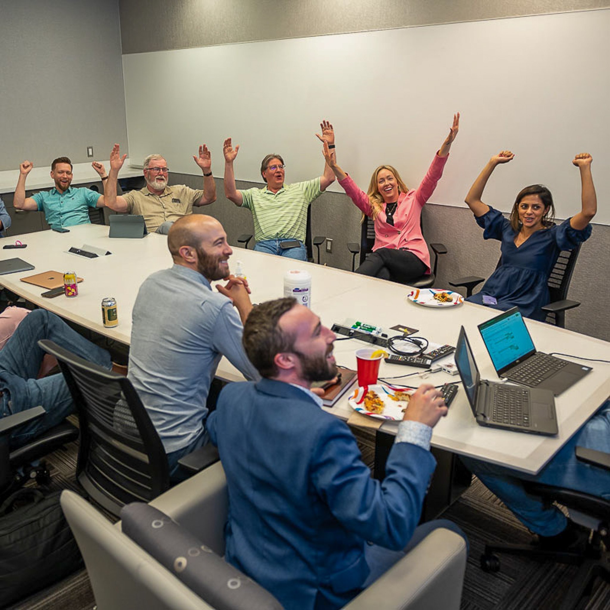 Group of people celebrating in a meeting room, some raising their arms and smiling, with food and drinks on the table.