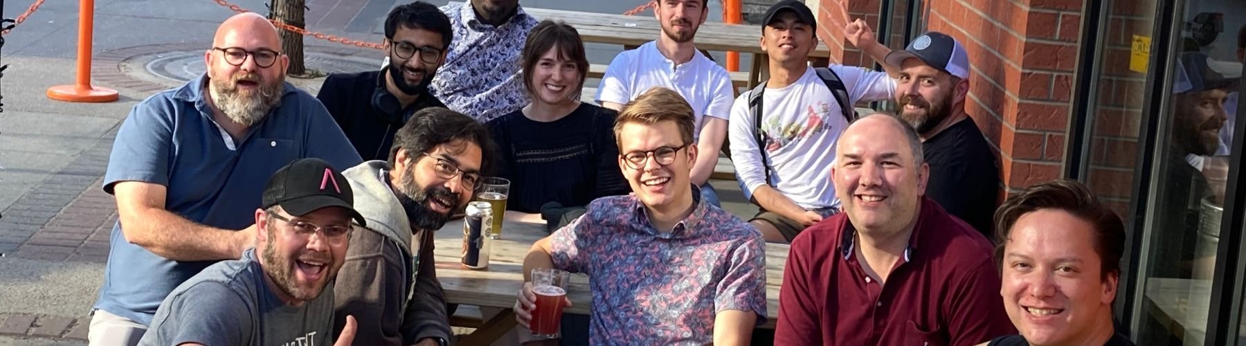 A group of people gathered outdoors at a wooden table, smiling and enjoying drinks in the sunlight.