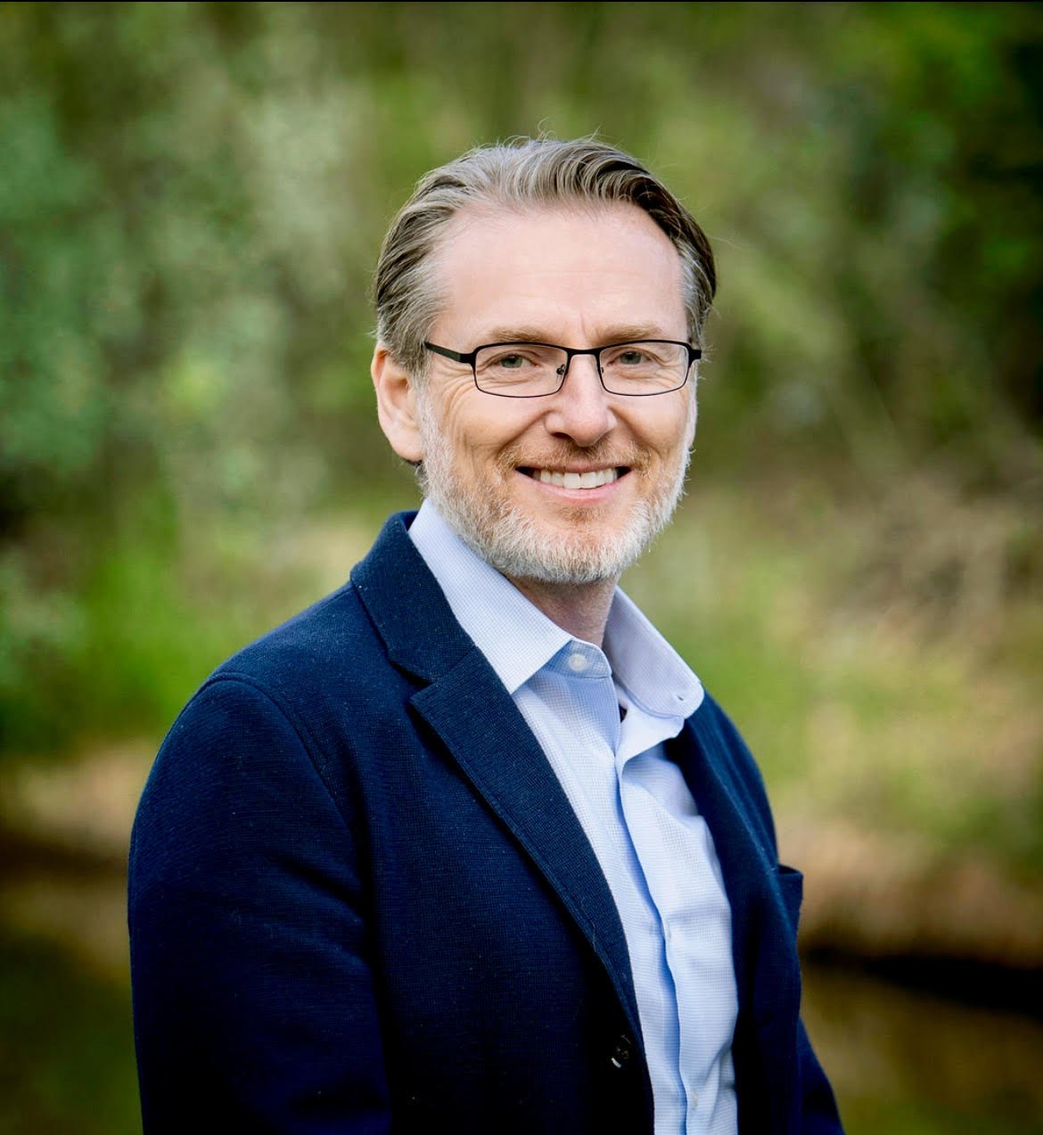 A smiling man with glasses, and a beard, wearing a blue blazer and light blue shirt, outdoors with a blurred green natural background.