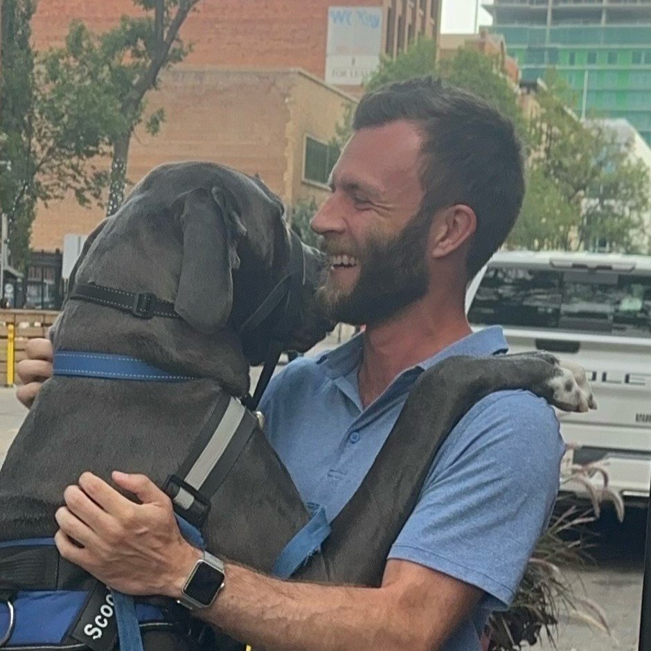 A man with a beard smiling and holding a large Great Dane dog in a blue harness, outside in an urban setting.