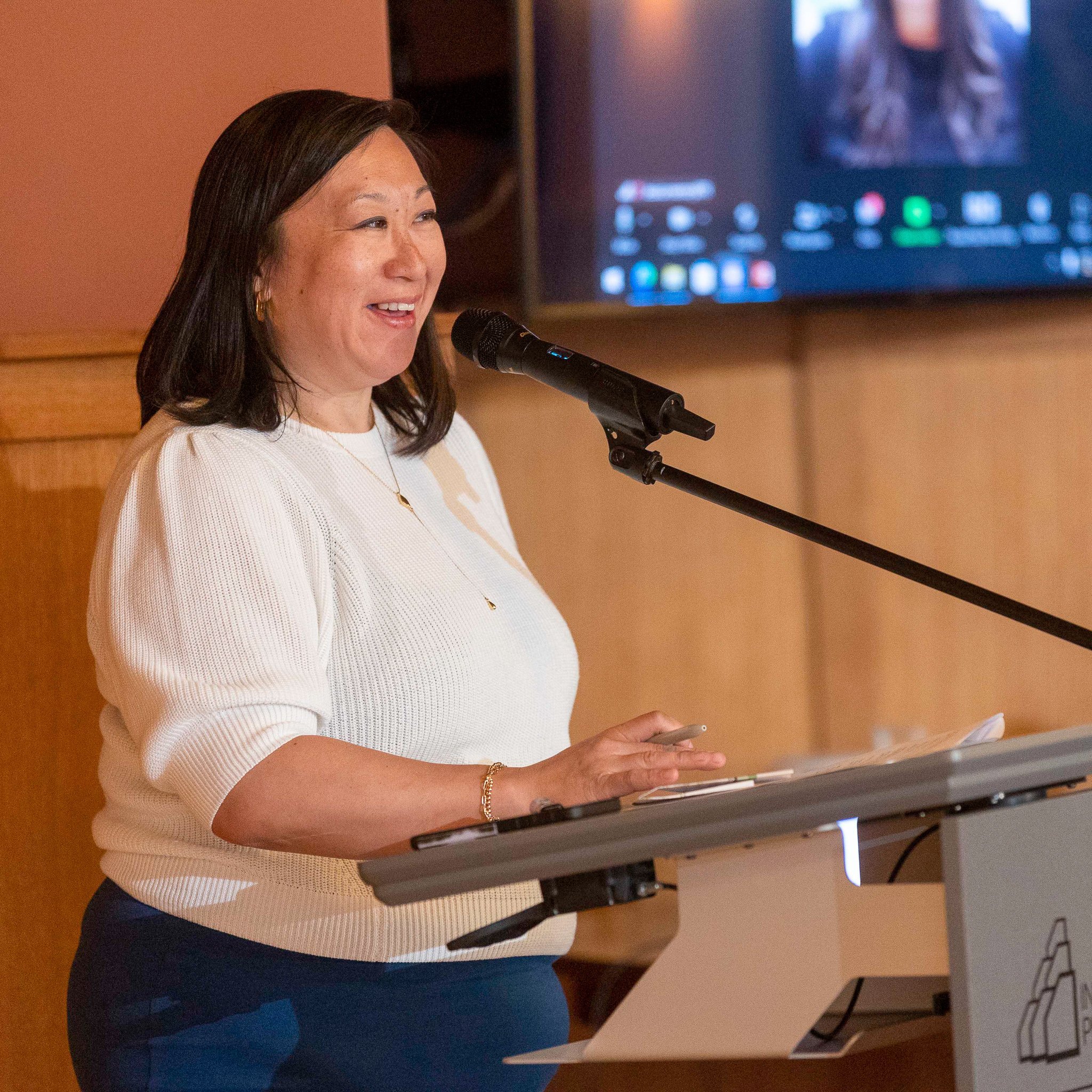 A woman speaking at a podium with a microphone in a room with wooden paneling and a large screen behind her displaying a video call interface.