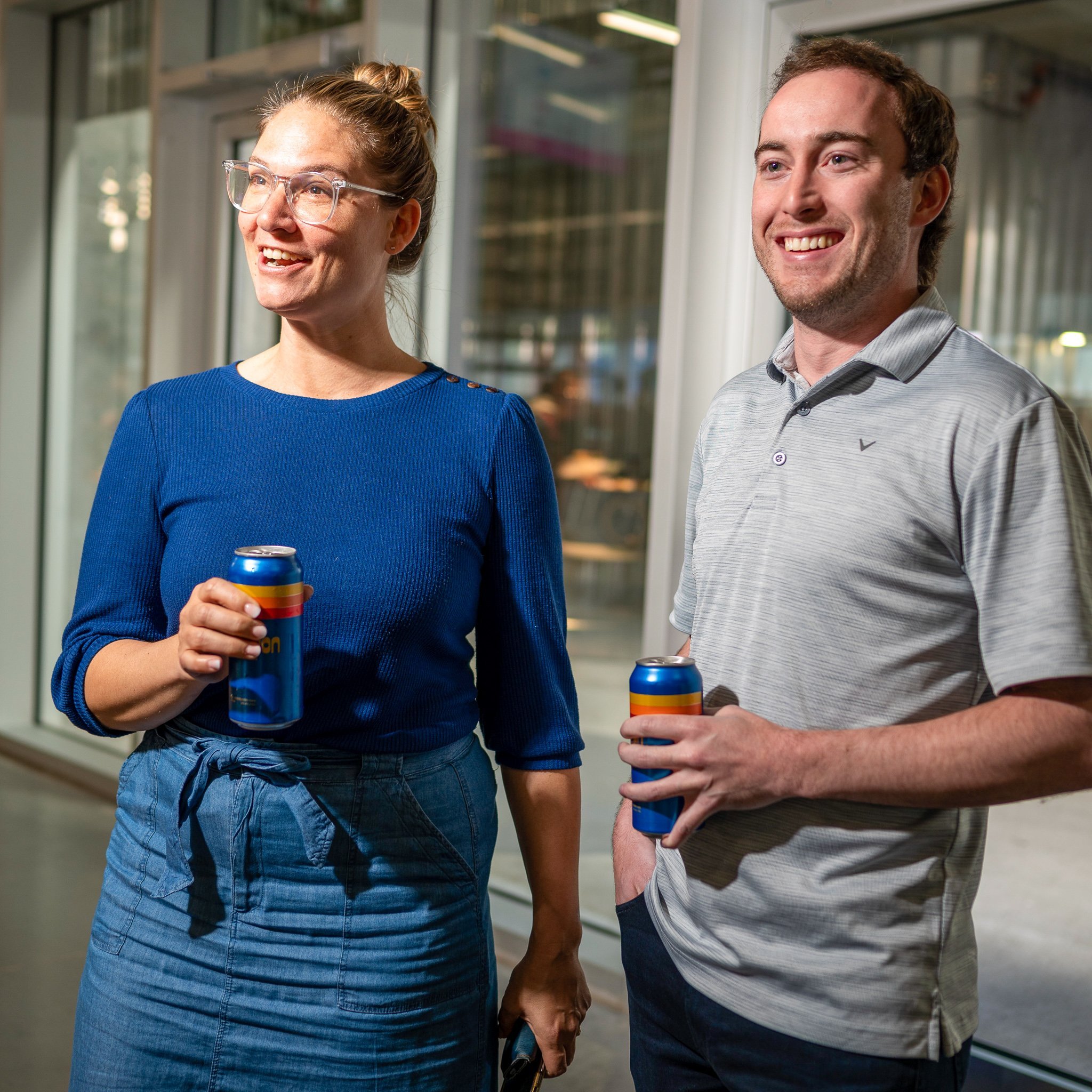 Two people, a woman and a man, standing indoors holding cans of soda and smiling, near a glass wall or window.