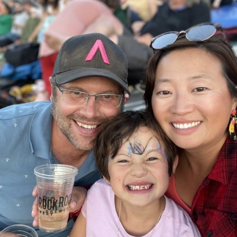 A smiling family of three taking a selfie at an outdoor event. The family includes an adult man wearing glasses and a black cap, an adult woman with sunglasses on her head, and a young girl with face paint and missing front teeth.