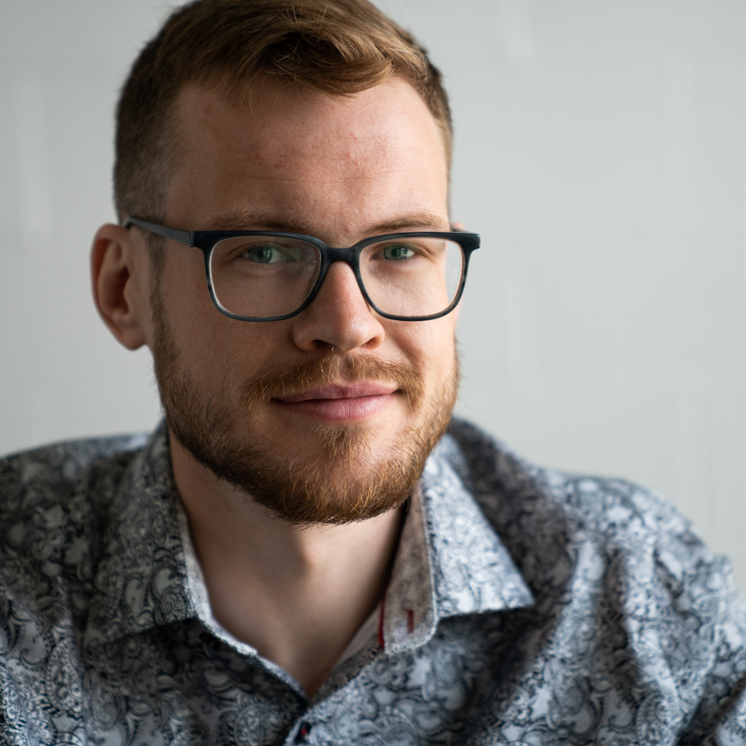 A man with red hair, glasses, and a beard, wearing a patterned shirt, smiling softly.